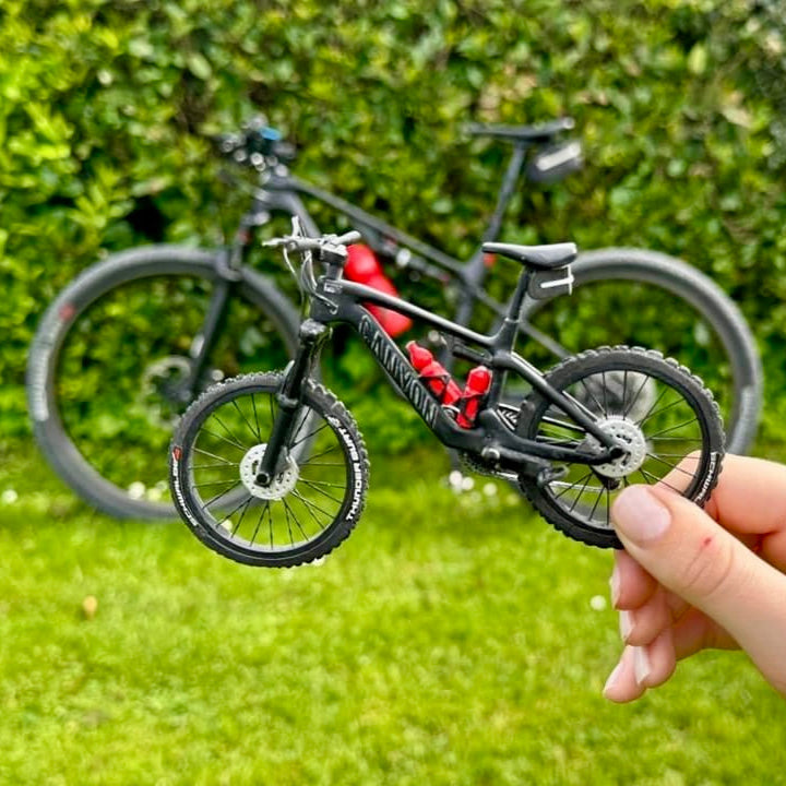 Hand holding a small model bicycle with a larger bicycle in the background.