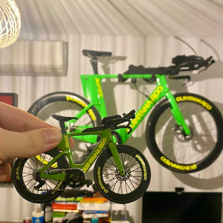 Green bicycle model held by a hand with a full-sized bicycle in the background.