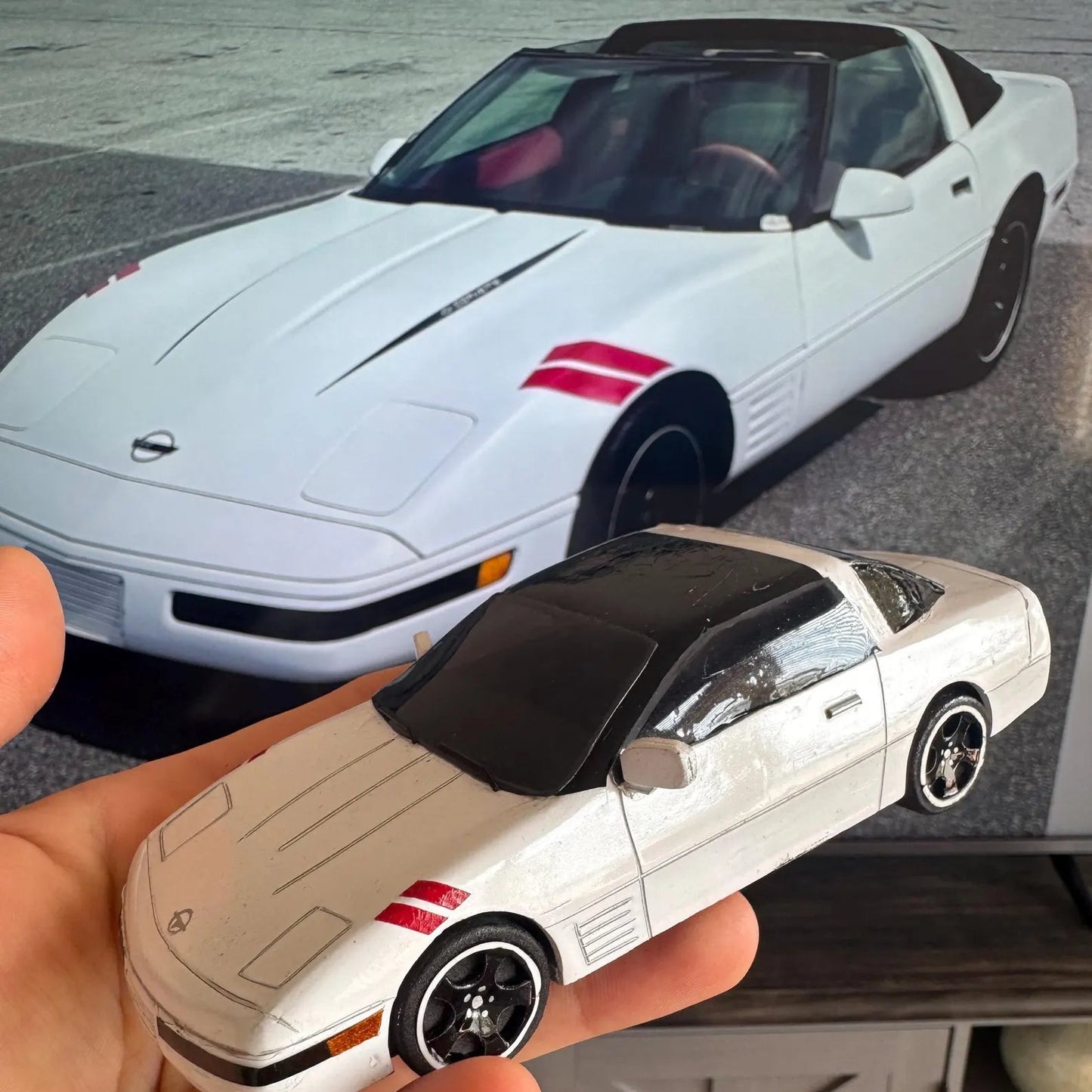 Hand holding a small model car in front of a larger white sports car.