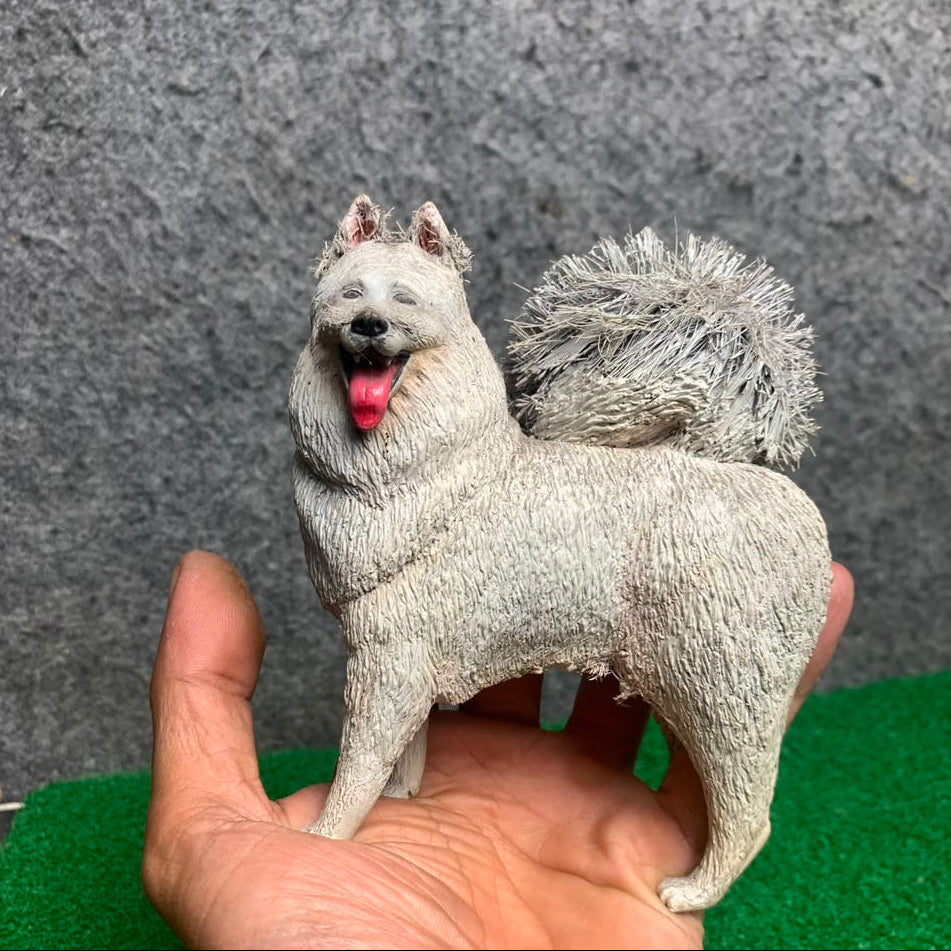 A small sculpted dog figurine with a fluffy gray-white coat and pink tongue, displayed on a person's hand against a textured gray wall and green surface.