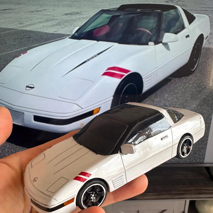 Hand holding a small model car in front of a larger white sports car.
